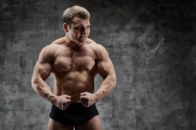 Sexy bodybuilder man posing on dark background in black shorts. Handsome pumped male body isolated with free space for advertising