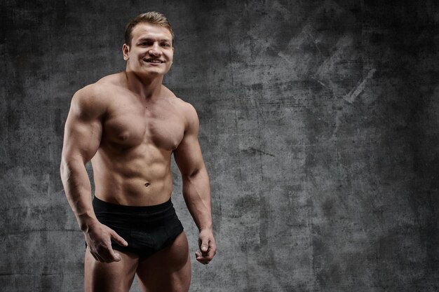 Sexy bodybuilder man posing on dark background in black shorts. Handsome pumped male body isolated with free space for advertising