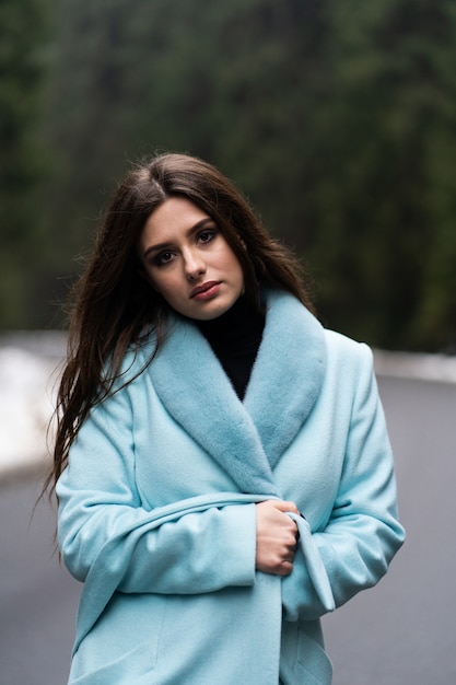 Sexy brunette girl poasing on road on winter mountains table. Glamorous young woman wearing stylish blue long coat. Fashion, business and beautiful people concept..