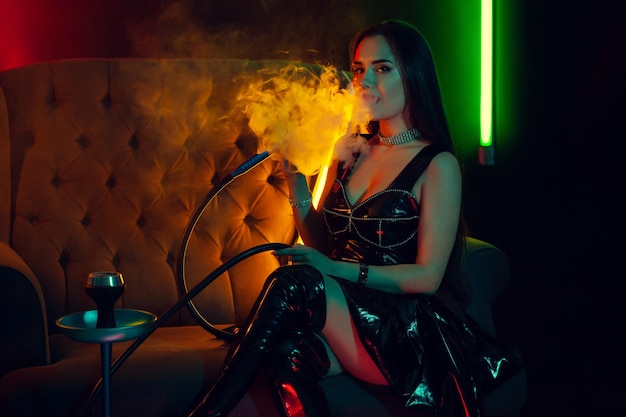 Sexy modella bruna sta fumando un narghilè esalando un fumo in un night club di lusso.