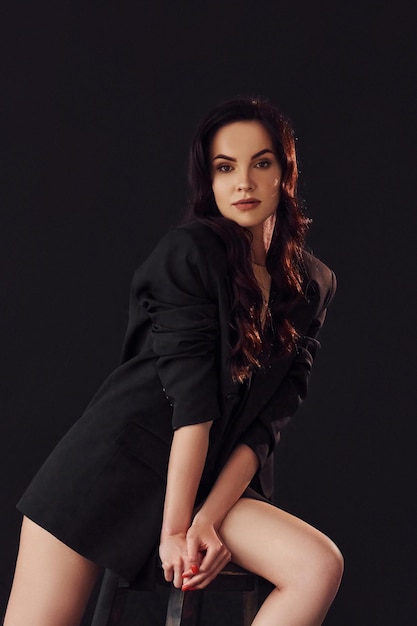 Sexy brunette with curly hair in luxury clothes sits on chair in the studio against black background