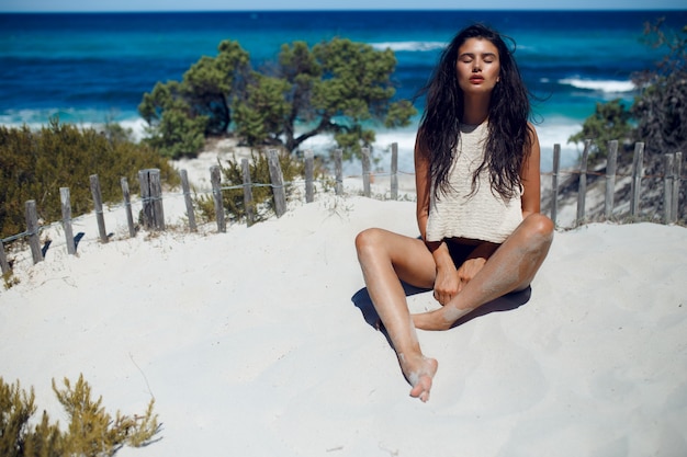 A sexy brunette woman with long hair, sitting on the hot sand on the beach with closed eyes, have relaxation time on Corsica island, sea waves and white beach background. Horizontal view.