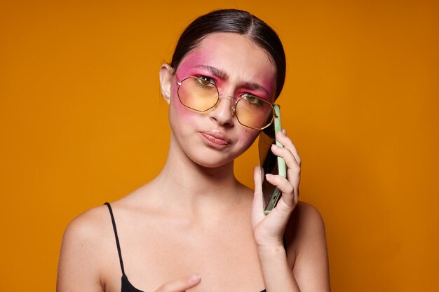 Сексуальная брюнетка с телефоном в розовом макияже лица позирует с привлекательным взглядом в очках на желтом фоне