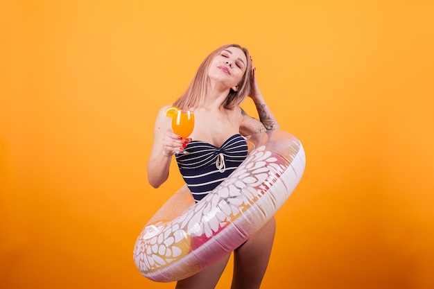 黄色の背景の上に膨脹可能な浮き輪を保持しているセクシーな白人女性。オレンジカクテルを持っています。水着姿の女性。