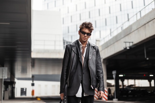 Sexy European young man in sunglasses in fashion youth oversized black leather jacket with a trendy hairstyle is resting near a modern building in the city on a spring day.Serious guy hipster outdoors