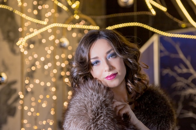 Sexy fashion woman wearing in winter fur coat. young woman in fur coat with red lips stands at the New Year's fair on the glitter garland background. Around the lights and festive mood.