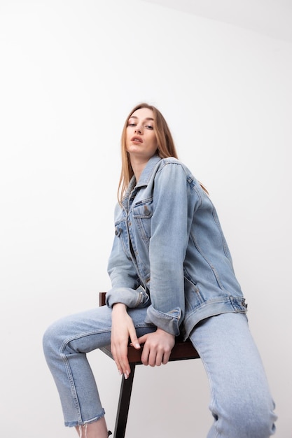 Sexy fashionable vogue woman model dressed in jeans and denim jacket sitting on chair white background