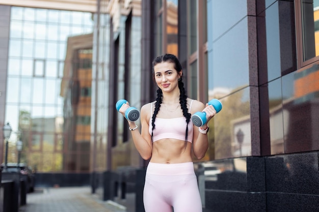 Sexy fit brunette woman with pigtails trains muscles with dumbbells in her hands on the background of a city building