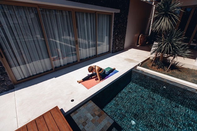 Sexy flexible and developed model wearing tight sportswear and doing yoga next to her villa and pool. Slender girl trying to sit on a twine on a yoga mat. Sports concept