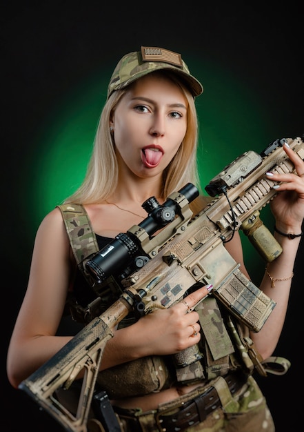 군용 에어소프트 작업복을 입은 섹시한 소녀가 어두운 배경에서 손에 총을 들고 포즈를 취하고 있습니다.