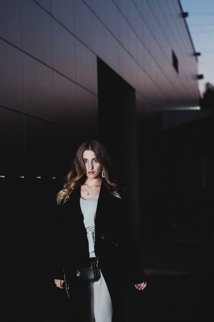 Photo sexy girl posing against a dark wall in the evening. an elegant fashionable girl in a long black coat, white jeans, with a leather bag stands near the wall of the building. beautiful girl for a walk