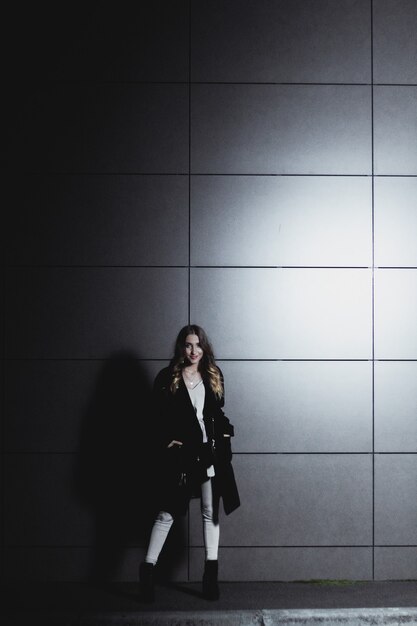 저녁에 어두운 벽에 포즈 섹시 한 여자. 긴 검은 코트, 흰색 청바지, 가죽 가방을 입은 우아한 유행 소녀가 건물 벽 근처에 서 있습니다. 산책을 위한 아름다운 소녀
