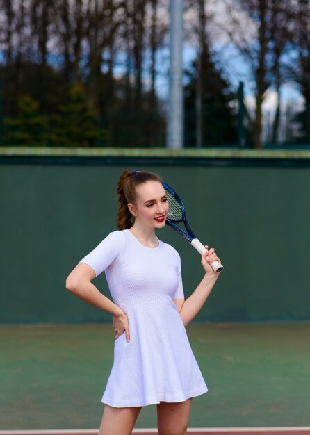 Tennis sexy della ragazza in vestito bianco e talloni che tengono la racchetta di tennis sulla corte.