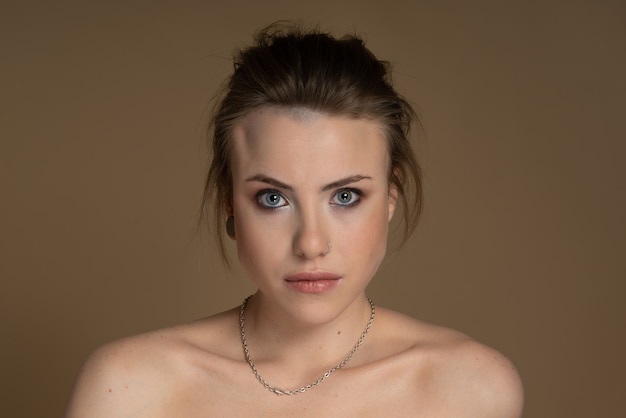 Sexy girl with heterochromia, looks expressively at camera, nose piercing and plug in one ear, and strange hairstyle. With amazing professional makeup and silver chain around her neck Beige background