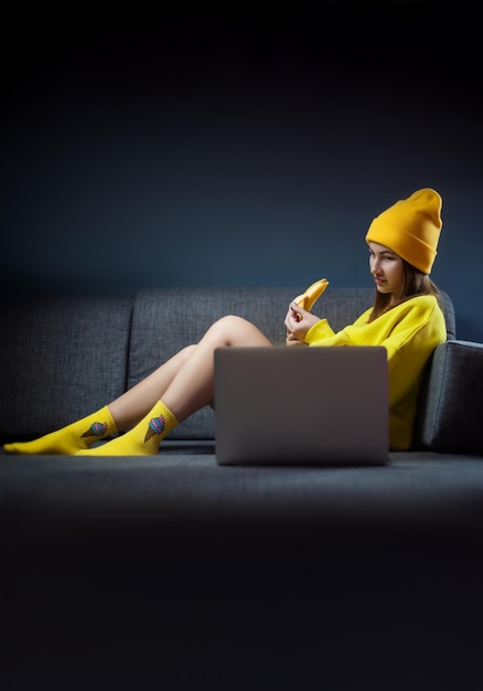 Сексуальная девушка в желтой одежде и шляпе с бананом и ноутбуком на темном фоне копирует пасту