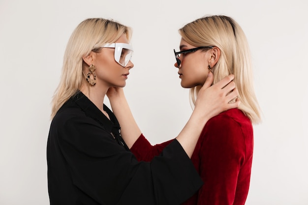 Sexy girlfriends in stylish dresses with trendy glasses look at each other and straighten their hair indoors