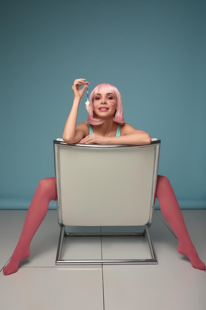 Сексуальная гламурная женщина с модным макияжем на красивом лице и короткой прической в розовом парике в платье ест мармеладную мышь в студии на сером фоне