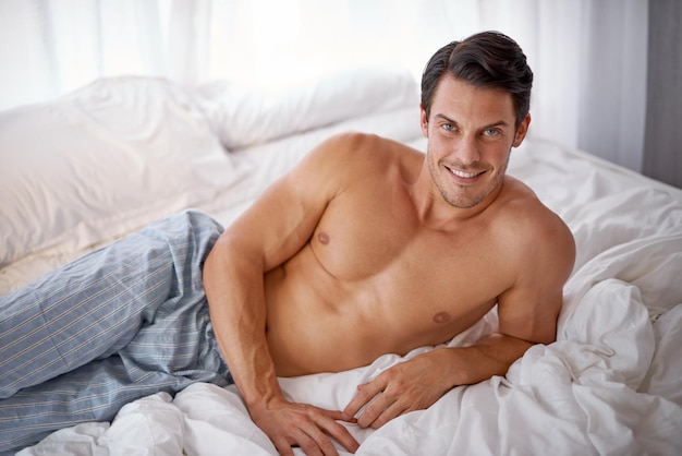 Сексуальный счастливый и портрет мужчины на кровати для расслабляющего комфорта и отдыха в пижаме Удобный уютный и красивый молодой человек, довольный в спальне, улыбающийся и уверенный, чтобы расслабиться на выходных