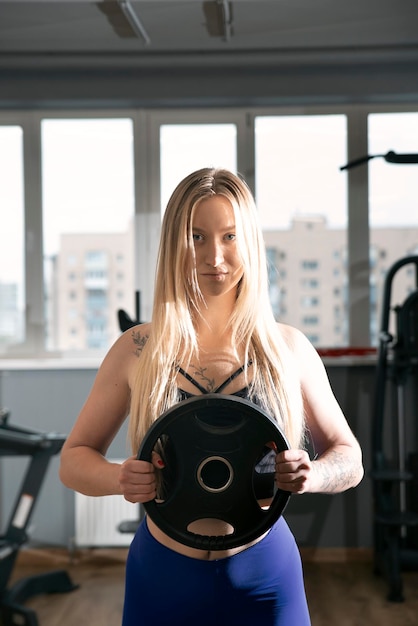 Sexy Kaukasische fitness model poseren in de sportschool. Blond meisje houdt halters. Sporten, gezonde levensstijl. Verticaal kader.