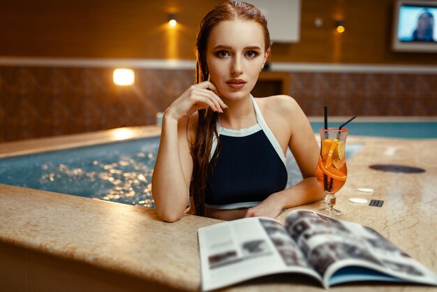 Sexy lady with fruit cocktail and journal at the pool side indoors.