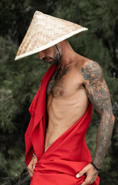 Sexy male monk in a red cape and asian hat in tattoos in nature