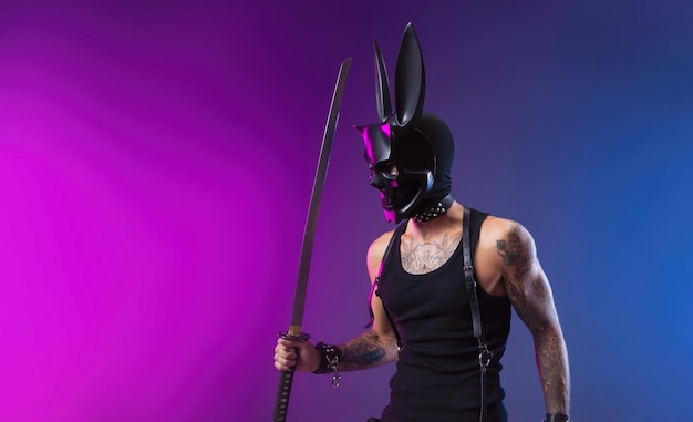 A sexy man in a demon mask and leather bdsm straps holds a katana sword in his hand on a neon background