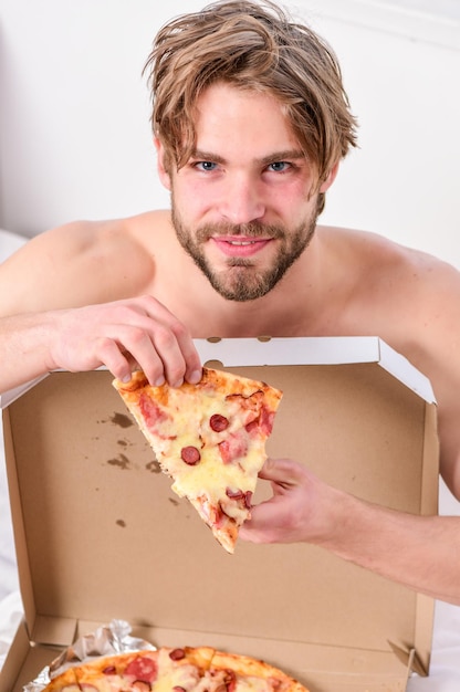 Sexy man eat pizza lying on bed Shirtless handsome young man with pizza on bed Hungry young man in