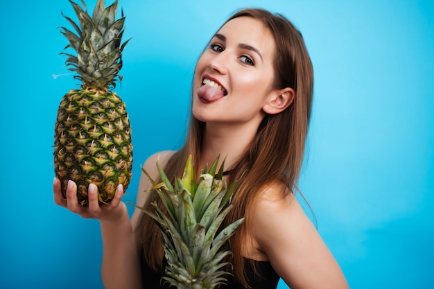 Sexy meisje in een badpak dat een rijpe ananas houdt die op blauw wordt geïsoleerd