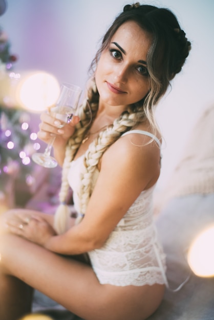 Sexy meisje in lingerie met twee staartjes en een glas champagne bij de kerstboom op het bed met een schattig plaid