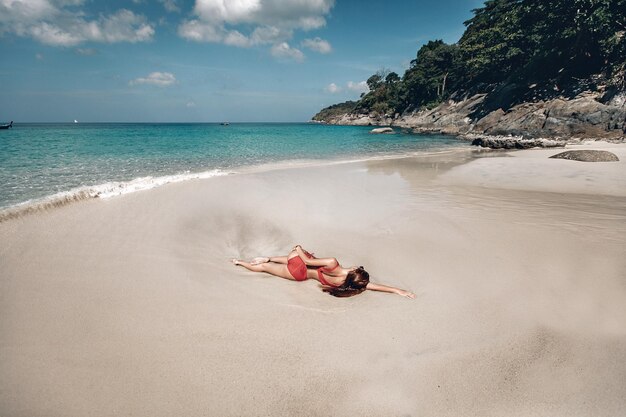 Sexy meisje in rode bikini zonnebaden op nat zand door de heldere oceaan, zonnige dag, rotsen, bomen; zonnebaden concept.