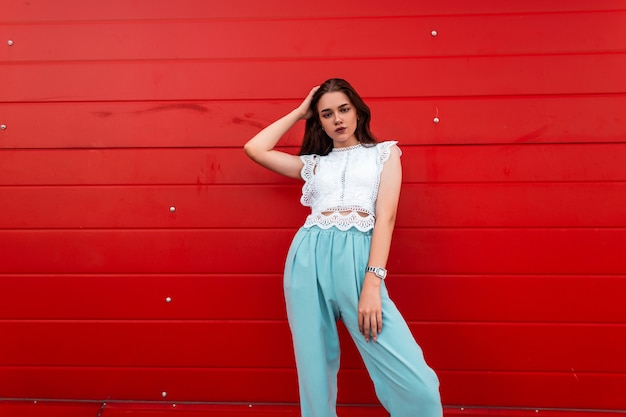 Sexy moderne jonge brunette vrouw in een elegante witte blouse in vintage blauwe broek rusten in de buurt van een rode houten muur in de stad.