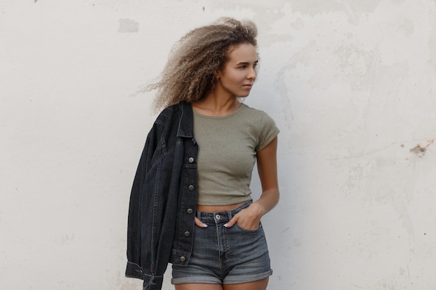 Sexy mooie jonge model vrouw met krullend haar in een t-shirt en jeans broek met jas poseren in de buurt van witte muur