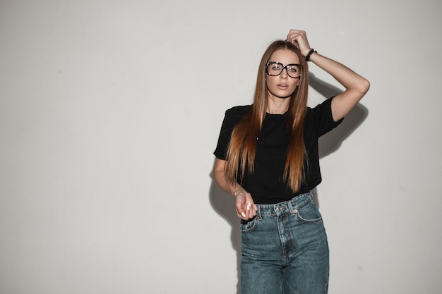 Sexy mooie vrouw model met vintage stijlvolle brillen bril in zwarte tshirt mockup en klassieke jeans poses in de buurt van witte muur mooi meisje in mode kleding in studio