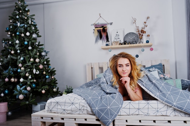 Sexy naked blonde model on bed with christmas gift boxes against new year tree.