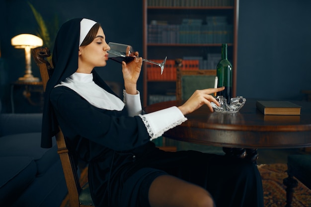 Sexy nun in a cassock sitting in a depraved pose with cigarette and glass of wine, vicious desires. Corrupt sister in the monastery, religion and faith, sinful religious people, attractive sinner