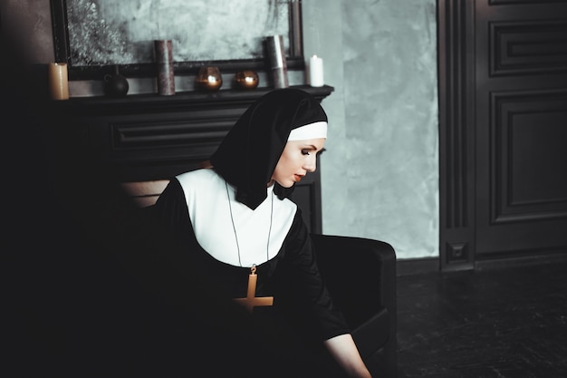 Photo sexy nun prays indoor . beautiful young holy sister. young beautiful nun with a cross in a robe in a black interior.