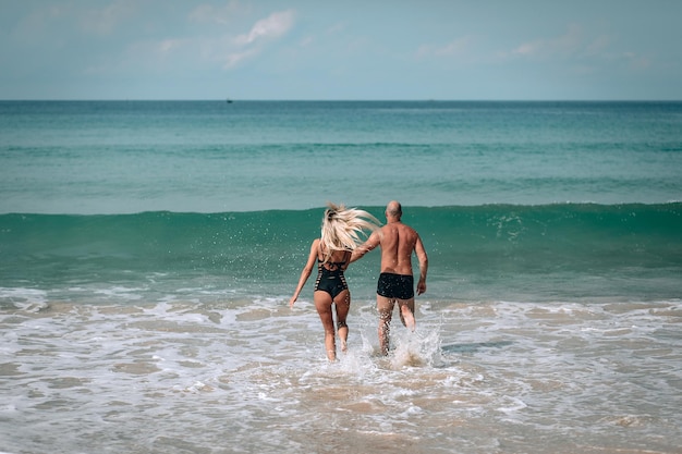 Sexy paar in het water. Grote golven van de zee. Mooie blonde in zwarte zwembroek met lang haar en een kale man zijn op vakantie. Achteraanzicht. Phuket. Thailand.