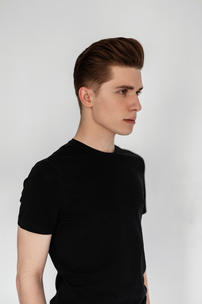 Sexy pretty young man in vintage vogue black t-shirt with fashionable hairstyle stands near white wall in room. Good-looking guy fashion model in stylish casual clothes indoors. Studio profile photo.