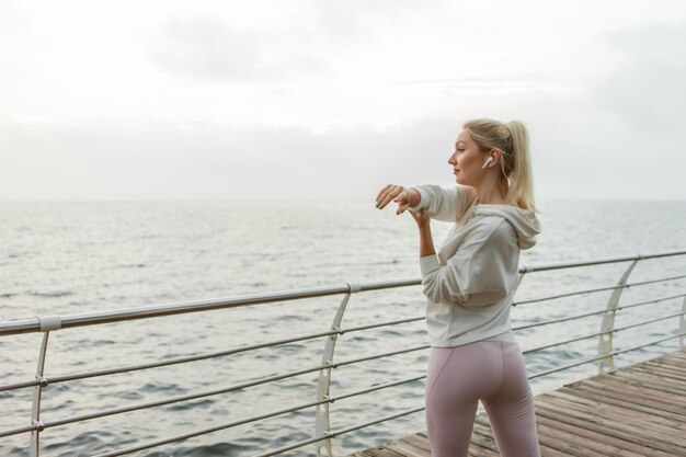 Sexy slim fit blonde vrouw oefent hand uitrekken voor outdoor training. Jong meisje gekleed in witte hoodie treinen op strand. Gezonde levensstijl, sport en fitness