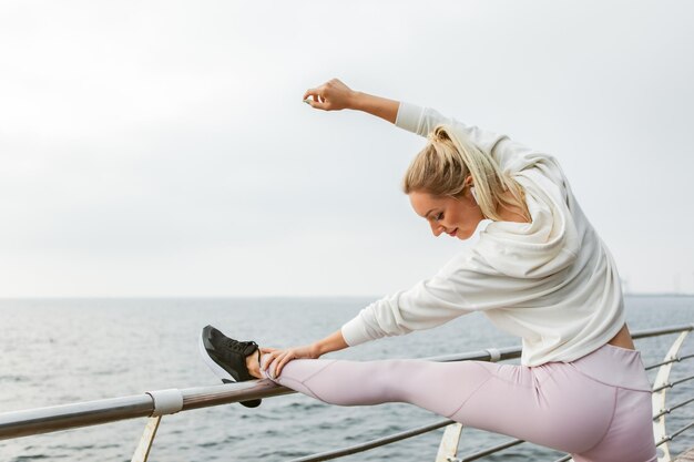 Sexy slim fit blonde woman is practicing leg stretching before outdoor workout. Young girl dressed in white hoodie trains on beach. Healthy lifestyle, sports and fitness