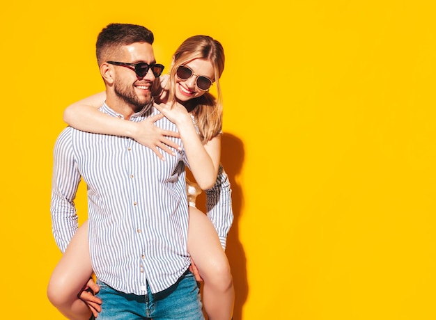 Sexy smiling beautiful woman and her handsome boyfriend Happy cheerful family having tender moments near yellow wall in studioPure cheerful models Gives piggyback ridingEmbracing in sunglasses