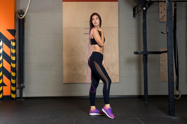 Sexy sports girl in black leggings with transparent fabric and in pink sneakers stands in the gym and holds a rope