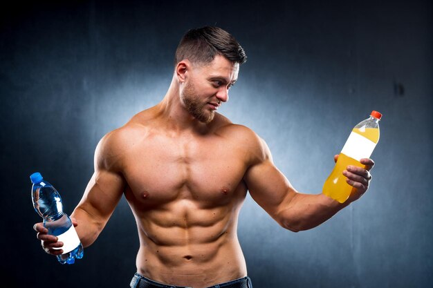 Sexy sportsman holding a bottle of water and soda choosing\
between healthy and harmful drink portrait looking at soda water\
closeup