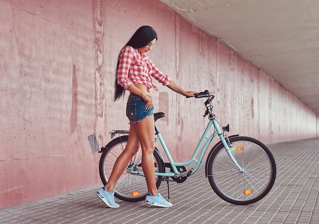 ピンクのフランネルシャツとメガネのデニムショートパンツを着て、ピンクの壁にシティバイクでポーズをとっているセクシーでスタイリッシュなブルネットの女の子。