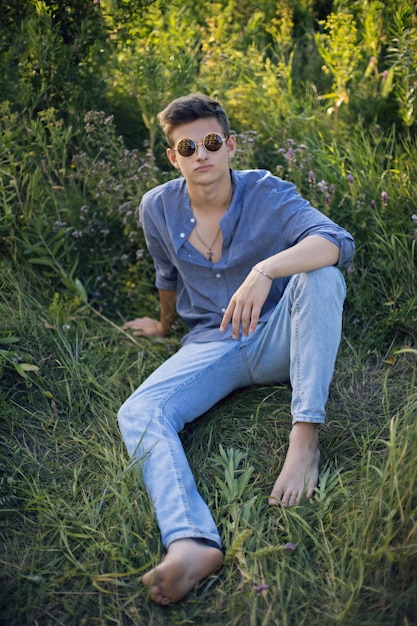 Sexy teen guy in sunglasses is sitting on the grass in nature in a shirt and jeans barefoot and looking at the camera