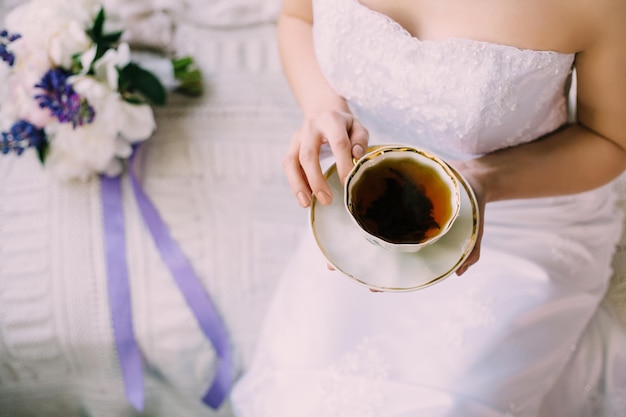 Sexy tender bride in night gown or wedding dress with decollete drinking a cup of morning black tea in porcelain cup and saucer sitting on the sofa Purple and white bouquet