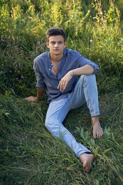 Sexy tienerjongen zittend op het gras in de natuur in een shirt en spijkerbroek op blote voeten en kijkend naar de camera