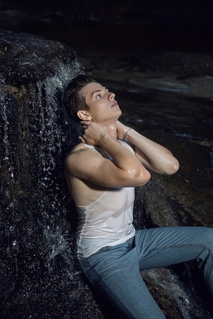 Сексуальный модный парень-подросток в белой футболке и синих джинсах сидит на скале у водопада в темном лесу летом