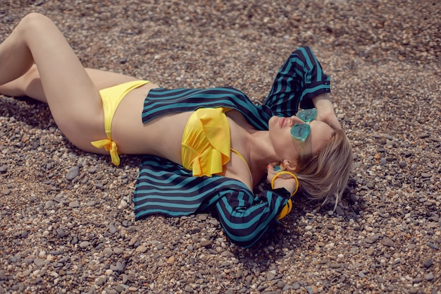 Sexy vrouw in een geel zwempak, een gestreept groen shirt en zonnebril ligt in de zomer op het strand aan zee