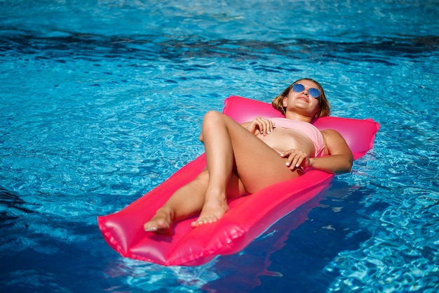 Sexy vrouw in een zwembroek ligt op een roze opblaasbare matras in het zwembad. Ontspan bij het zwembad op een warme zonnige zomerdag. Vakantie concept